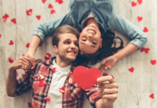 Top view of happy young couple looking at camera and smiling while lying on wooden floor. Guy is holding a red paper heart