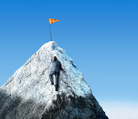 A businessman climbs a steep snow capped mountain peak in order to reach a flag that is planted at the top.