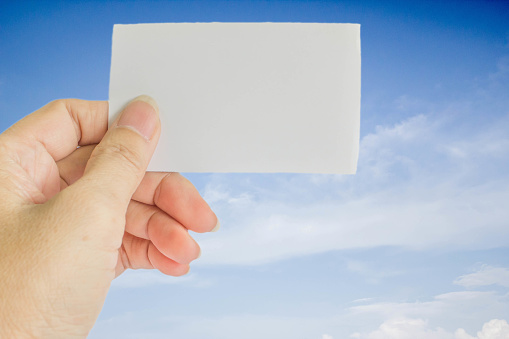concept photo of left hand hold business card, credit card or blank paper on blurrd blue sky with white cloud
