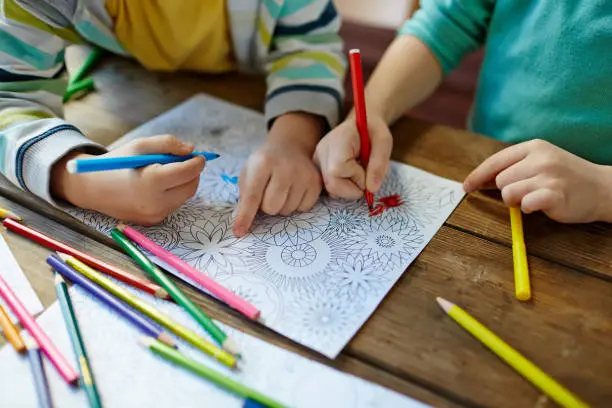 Close-up shot of two pairs of little hands coloring mandala with felt-tip pens; pencils of different colors and illustrations laid on wooden table