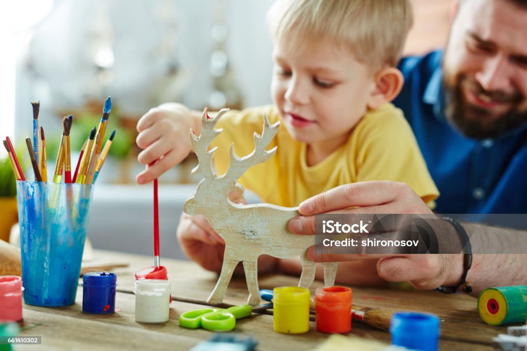 Handmade Christmas present for granny Little blond-haired son with his bearded dad preparing Christmas present for granny: they sitting at table and enthusiastically coloring toy deer in different colors Child Stock Photo