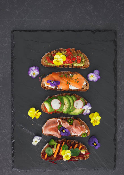 Spring tea sandwiches with flowers and herbs Rye bread sandwiches with avocado and cheese cream, prosciutto and salmon fish, edible flower garnish. Top view, blank space edible flower stock pictures, royalty-free photos & images