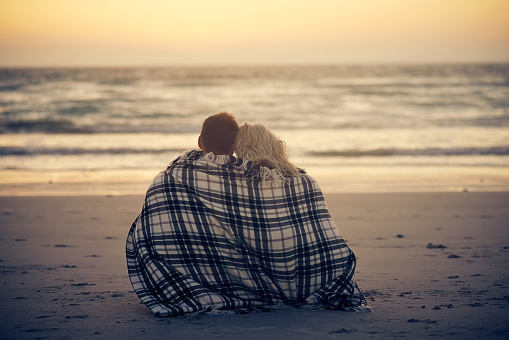 Rearview shot of a young couple sitting on the beach with a blanket wrapped around them
