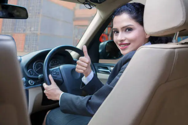 Successful Indian businesswoman driving a car and showing thumb up with container background on the car window