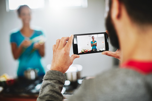 Shot of a man filming his wife on his smartphone while she cooks in their kitchen at home