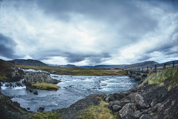 Mountain river in the severe gloomy Icelandic landscape. Stones and moss in the foreground. The picture was taken in cloudy cloudy weather in the autumn afternoon. The picture shows a bridge across the river