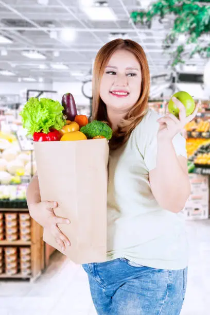 Overweight woman with blonde hair, holding a green apple fruit and shopping bag full of vegetables in the supermarket