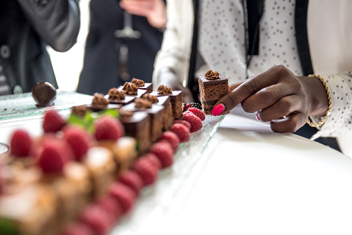 African woman's hand taking a small chocolate cake from the glass plate with chocolate cakes and raspberries on a white bar table.A woman has pink nail polish and she is wearing white dotted blouse and white jacket. Some men in the backrgound.