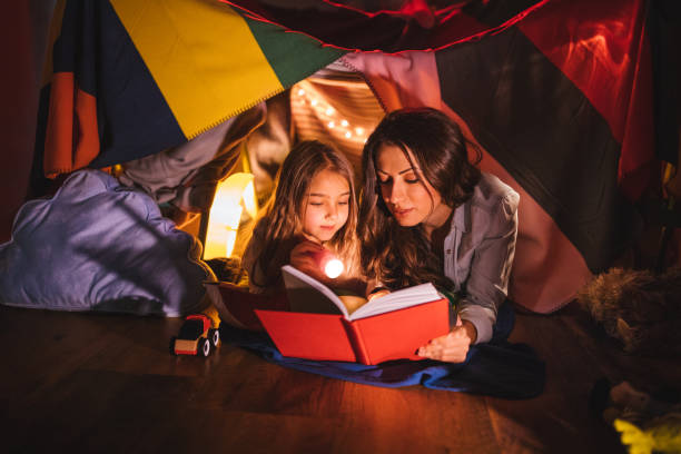 Mother reading a book to her daughter at night Mother reading a fairy tale book to her daughter under children's blanket fort at night fort stock pictures, royalty-free photos & images