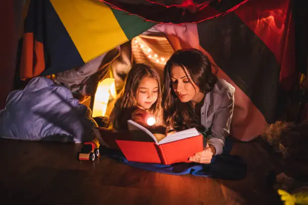 Mother reading a fairy tale book to her daughter under children's blanket fort at night