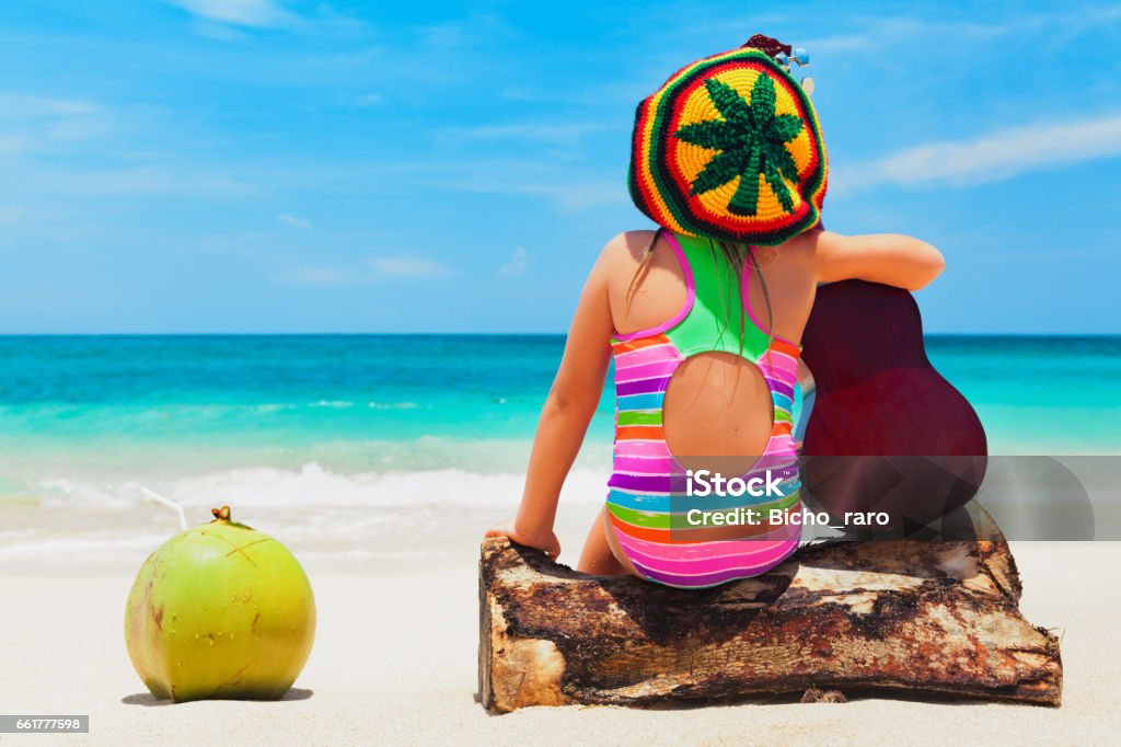 Happy baby have fun on summer tropical beach holiday Little happy baby girl in rastaman hat has fun, play reggae music on Hawaiian guitar, enjoy caribbean beach party. Children healthy lifestyle. Travel, family activity on Jamaica island summer holiday Beach Stock Photo