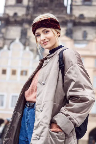 Portrait of a beautiful blonde woman in a cold winter in the historic district. She's wearing a warm winter jacket, pink sweater, jeans, backpack and ears are covered by headband. Captured are also falling snowflakes.