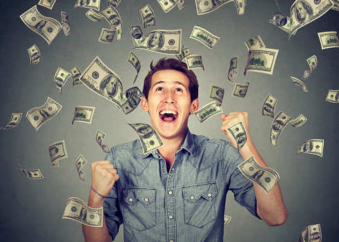 Happy young man screaming super excited. Portrait ecstatic guy celebrates success under money rain falling down dollar bills banknotes isolated gray background. Financial freedom concept