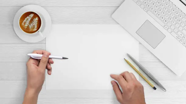 Direct view at hands writing on a blank paper . Office desk with laptop, coffee, notebook, pencils, and a plant. vintage wood white desk. copy space. hipster template