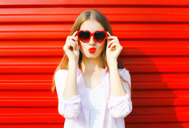 Portrait pretty young woman in red heart shape sunglasses over colorful background blowing lips Portrait pretty young woman in red heart shape sunglasses over colorful background blowing lips blowing a kiss stock pictures, royalty-free photos & images