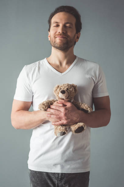 Handsome man with a toy Handsome man is holding a teddy bear and smiling with closed eyes, on gray background man with teddy bear stock pictures, royalty-free photos & images