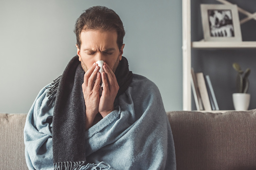 Handsome sick man is wiping nose while sitting on couch covered in plaid