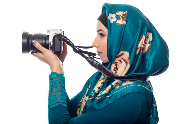 Female photographer wearing a hijab.  She is either a hobbyist or a journalist.  The headscarf is associated with muslims and east european or middle eastern culture.  she is holding a camera.