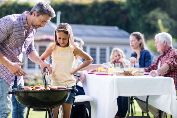 Father and daughter at barbecue grill while family having lunch in background Father and daughter at barbecue grill while family having lunch in the garden south african braai stock pictures, royalty-free photos & images