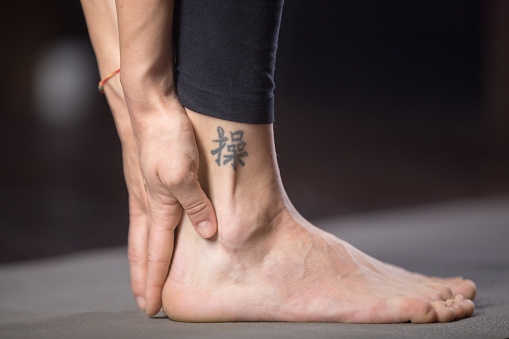 Woman practicing yoga, standing in forward bend exercise, head to knees, uttanasana, pose, working out, studio evening practice, close up, with tattoo on leg meaning Patience in studying