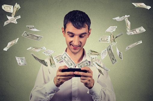 Technology online banking money transfer, e-commerce concept. Happy young man using smartphone with dollar bills flying away from screen isolated on gray wall office background.