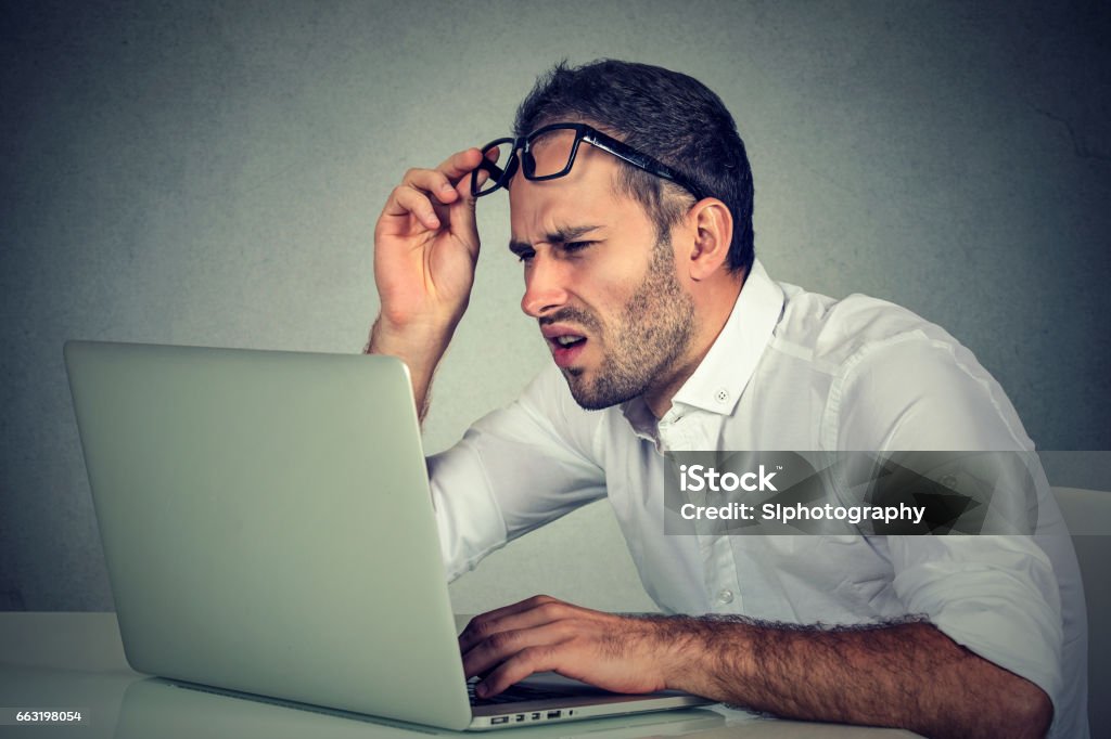 business man with glasses having eyesight problems Closeup portrait business man with glasses having eyesight problems confused with laptop software isolated on gray background. Vision related changes. Human face expression Confusion Stock Photo