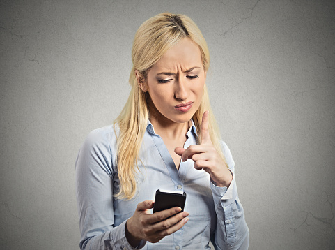 Portrait angry young business woman, mad displeased wife, pissed off employee holding mobile pointing with finger at smartphone isolated grey wall background. Negative human emotion facial expression