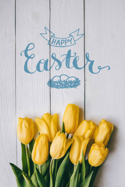 istock Happy Easter hand lettering calligraphy text. Yellow tulips bunch on white wooden planks rustic barn rural table background. Letters, inscription. Beautiful vertical flat lay postcard. 663781626