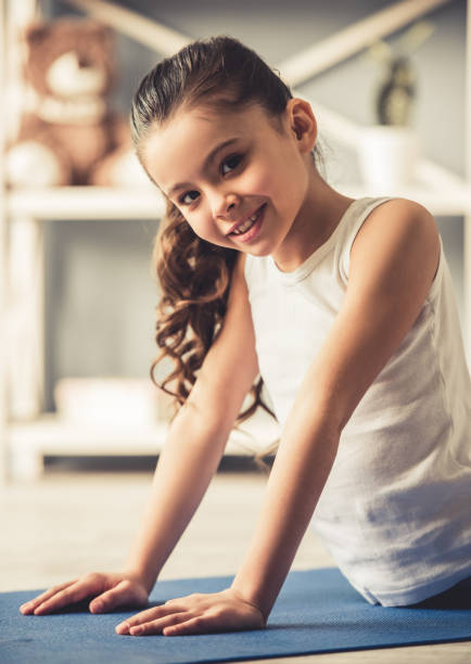 Charming little girl is looking at camera and smiling while doing yoga at home