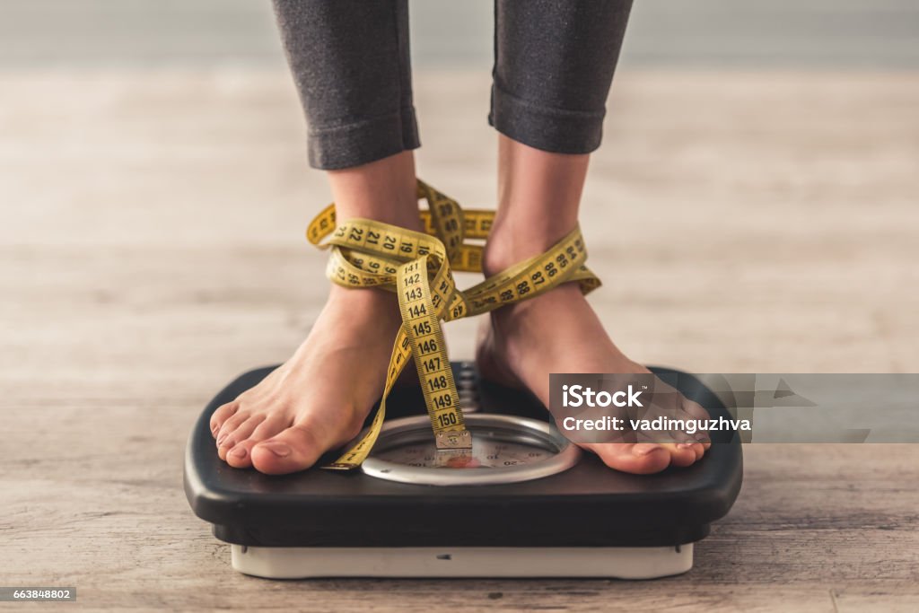 Girl and weight loss Cropped image of woman feet standing on weigh scales, on gray background. Legs winded with a tape measure Dieting Stock Photo