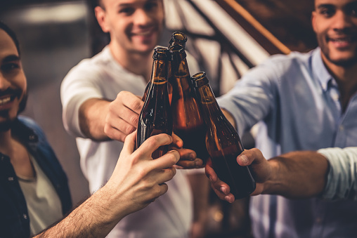 Handsome guys are clinking bottles of beer and smiling while resting in pub