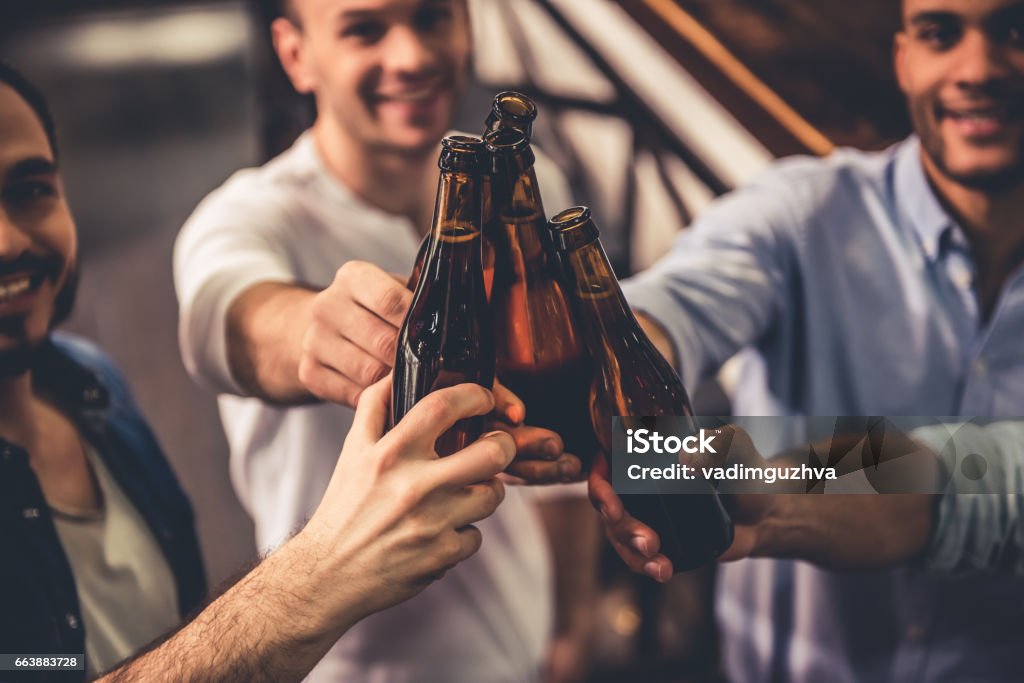 Friends in pub Handsome guys are clinking bottles of beer and smiling while resting in pub Beer - Alcohol Stock Photo