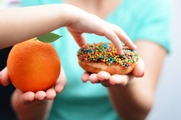 Child obesity concept with little girl hand choosing a sweet and unhealthy doughnut instead of a fruit stock photo