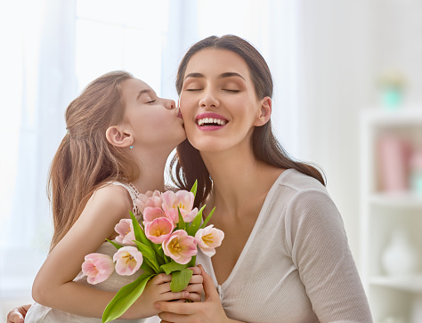 Happy mother's day! Child daughter congratulates mom and gives her flowers tulips. Mum and girl smiling and hugging. Family holiday and togetherness.