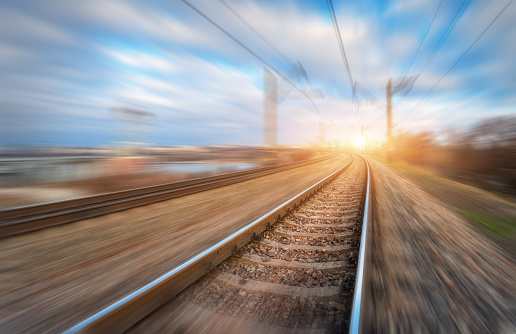 Railroad in motion at sunset. Railway station with motion blur effect and colorful sky with clouds. Industrial concept background. Railroad travel, railway tourism. Blurred railway. Transportation