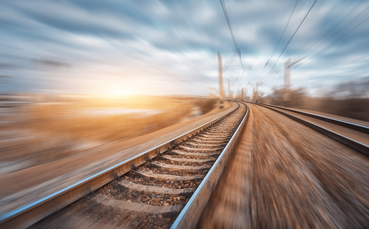 Railroad in motion at sunset. Railway station with motion blur effect and colorful sky with clouds. Industrial concept background. Railroad travel, railway tourism. Blurred railway. Transportation