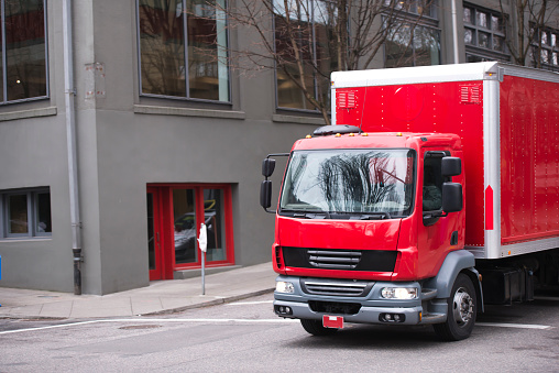 A bright red little buggy semi truck with a covered trailer box for transporting small loads and delivering goods to local stores and companies rides down the street of a modern urban city