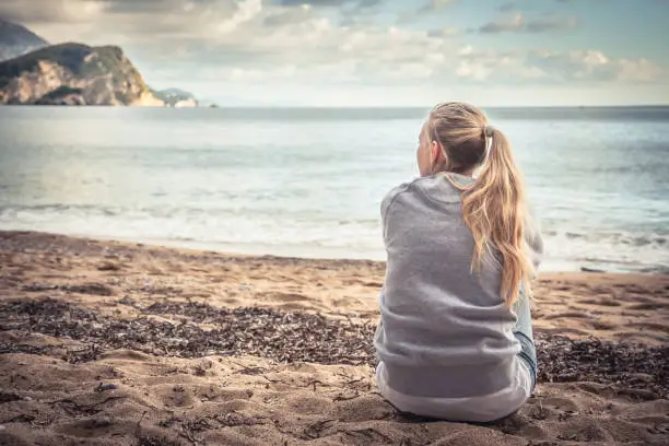 Photo of Pensive lonely young woman sitting on beach hugging her knees and looking into the distance with hope