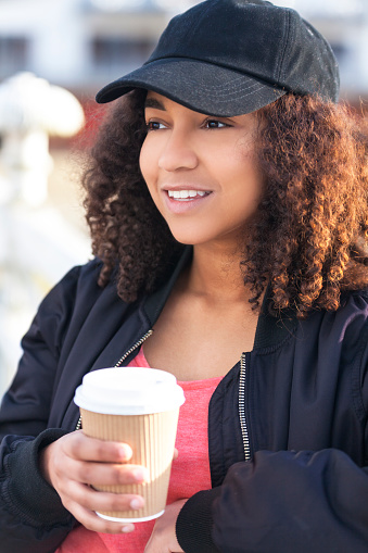 Beautiful happy mixed race African American girl teenager female young woman smiling drinking takeaway coffee outside wearing black baseball cap and bomber jacket