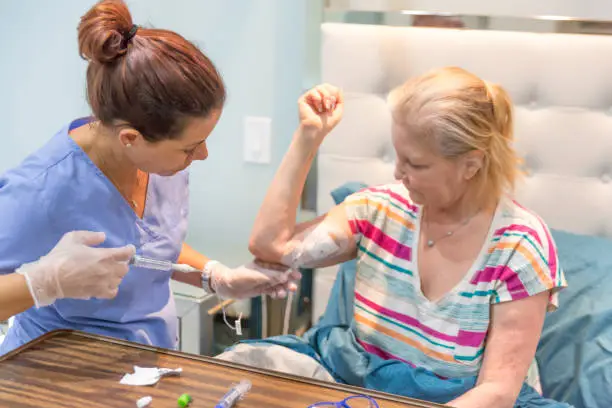 Middle aged female patient sits in bed in her home as nurse flushes out her picc line. Shot with Canon 5D Mark lll.