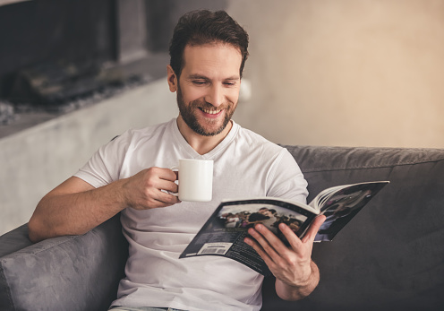 Handsome man is reading a magazine, holding a cup and smiling while resting at home