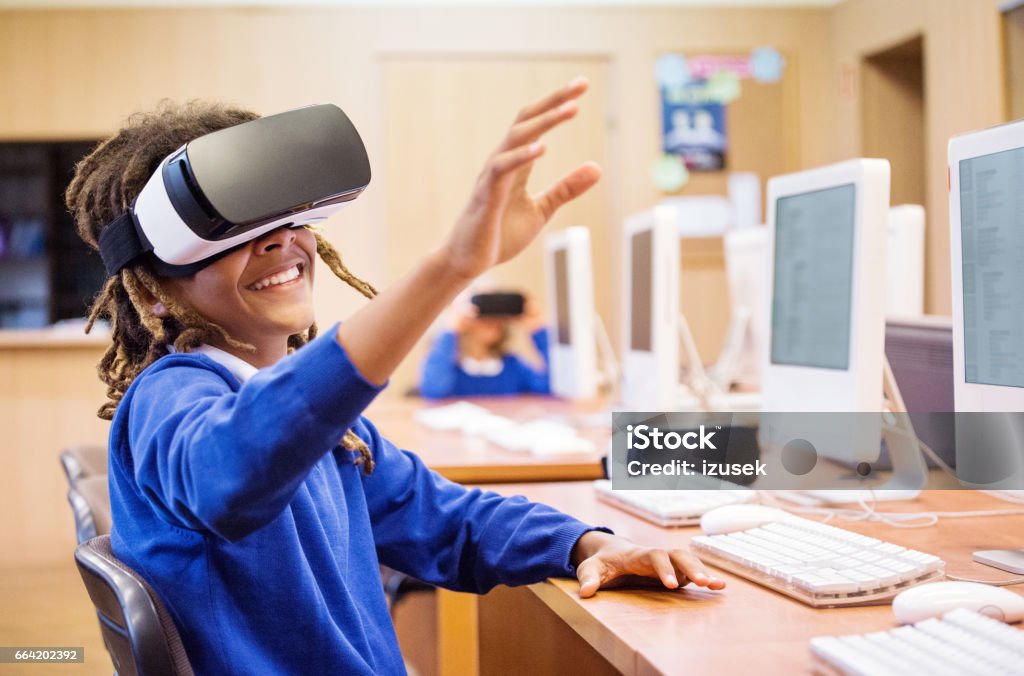Afro american student using virtual reality goggle at school Afro american student sitting in a computer lab at school and using virtual reality goggle, raising hand. Virtual Reality Simulator Stock Photo