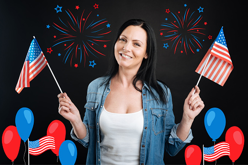 American holiday. Attractive delighted brunette woman holding US flags and enjoying herself while celebrating Independence Day