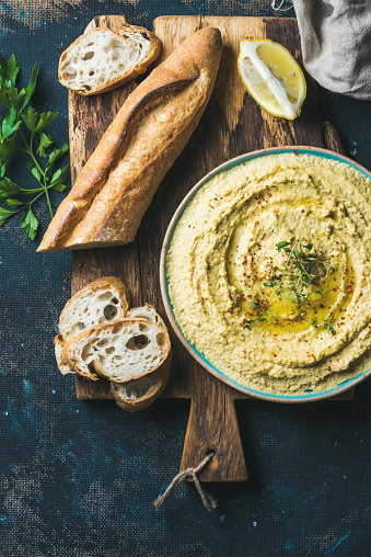 Homemade hummus with lemon, herbs and baguette on rustic wooden serving board over dark blue plywood background, top view, vertical composition, Vegetarian food concept