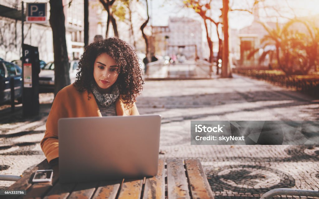 Beautiful curly lady with laptop in street bar Attractive beautiful adult brunette freelancer girl with black curly hair sitting in street cafe and working on her laptop with copy space for young advertising text message or promotional content Remote Location Stock Photo