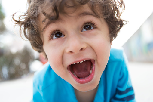 A close up of a young boy shouting or singing with his mouth wide open.  He is smiling and looking up, away from the camera.  He is wearing a blue and gray shirt, has brown hair, and is of Persian descent.  Photographed outdoors.