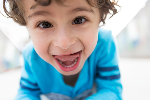 A close up of a young boy making a goofy face with his mouth wide open and his tongue out.  He is smiling and looking at the camera.  He is wearing a blue and gray shirt, has brown hair, and is of Persian descent.  Photographed outdoors.