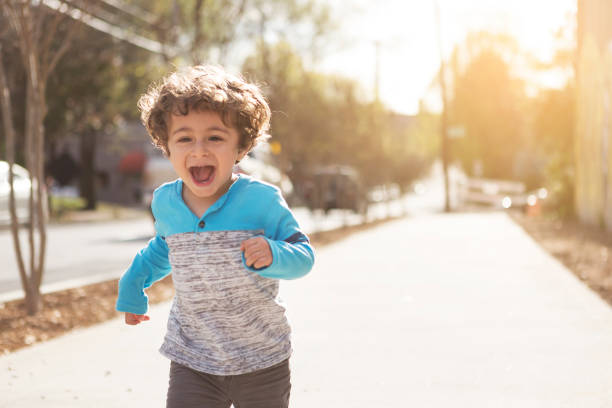 A  young boy running down the sidewalk.  He is smiling and yelling.  He is wearing a blue and gray shirt, has brown hair, and is of Persian descent.  Photographed outdoors, behind the subject is a street with parked cars and some homes.