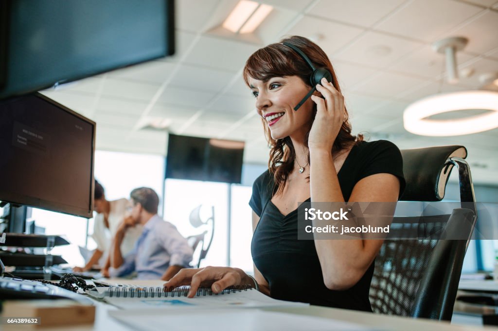 Beautiful mature business woman talking on headset Beautiful mature business woman talking on headset while working at her desk. Caucasian female executive with people working in background. Headset Stock Photo