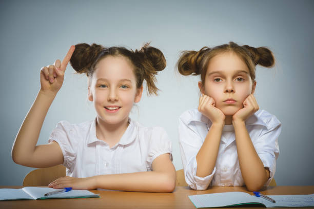 Thoughtful girl and happy girl sitting at desk on gray. school concept stock photo
